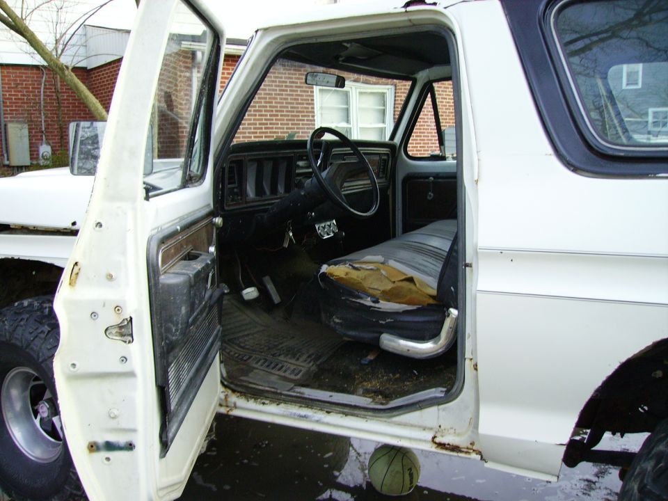 White 1978 Ford Bronco interior with torn driver's seat, exposed foam, and black dashboard. The driver's door is open
