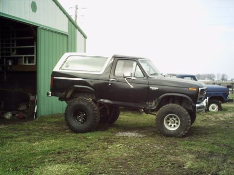 1986 Ford Bronco - A lifted black Ford Bronco sits in a grassy field next to a blue pickup truck and a green barn.