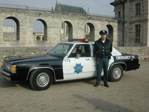 1991 Ford Crown Victoria - A police officer stands beside a black and white patrol car with 'EMERGENCY 911' on the rear quarter panel.