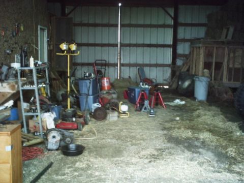 1986 Ford Bronco - A cluttered barn interior contains various tools, equipment, and hay, with a large metal door at the back.