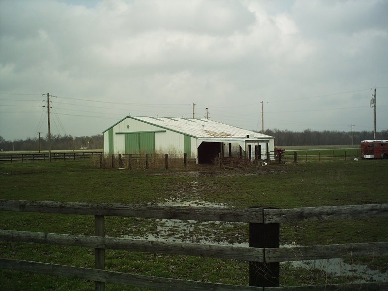 Barn with green doors and a red trailer in a muddy field under a cloudy sky