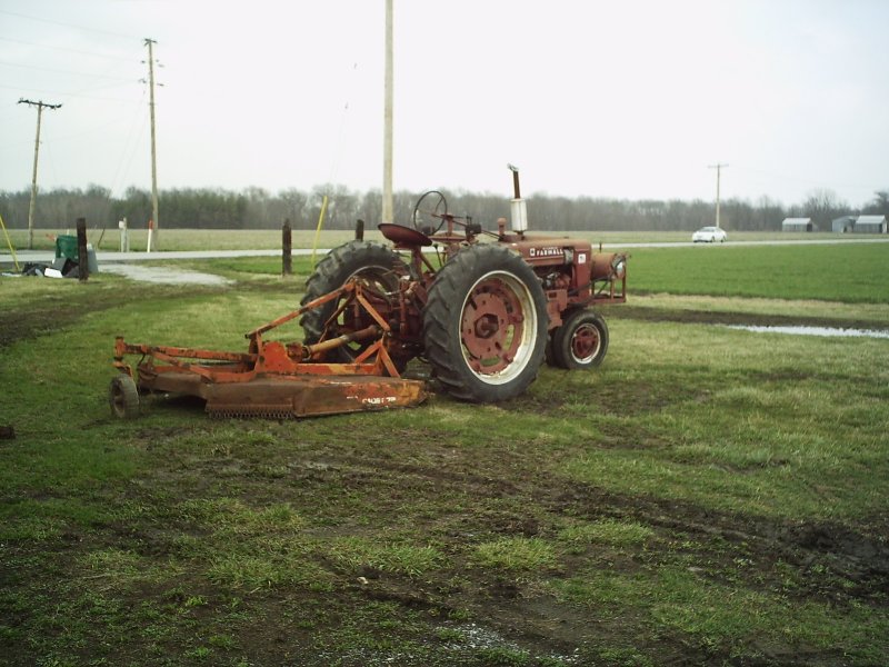 Red tractor with a rotary mower attached, parked on a muddy field. A white car is visible in the distance on a road