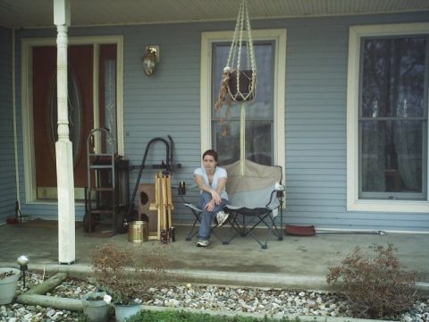 1986 Ford Bronco - A woman sits on a porch, surrounded by household items and plants, with a house in the background.