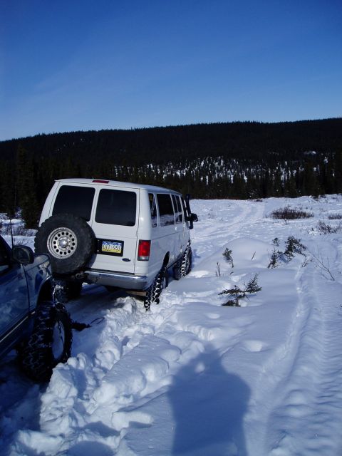 2000 Ford E350 - White Ford E350 van with spare tire on back stuck in deep snow next to another vehicle.
