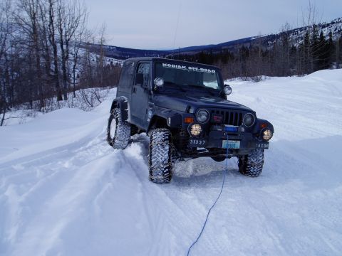 2000 Ford E350 - A dark blue Jeep Wrangler navigates a snowy terrain, equipped with off-road tires and auxiliary lights.