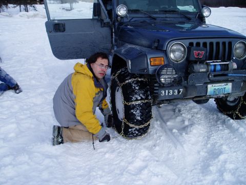 2000 Ford E350 - A person installs snow chains on the tire of a dark blue Jeep Wrangler in a snowy environment.