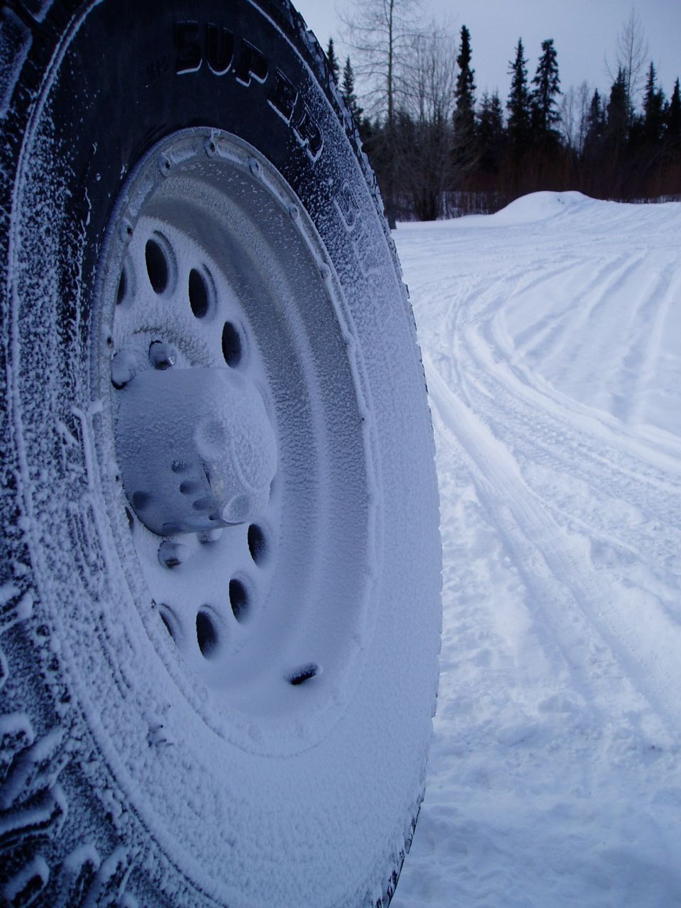 Close-up of a snow-covered tire and wheel in a snowy outdoor setting with tire tracks visible