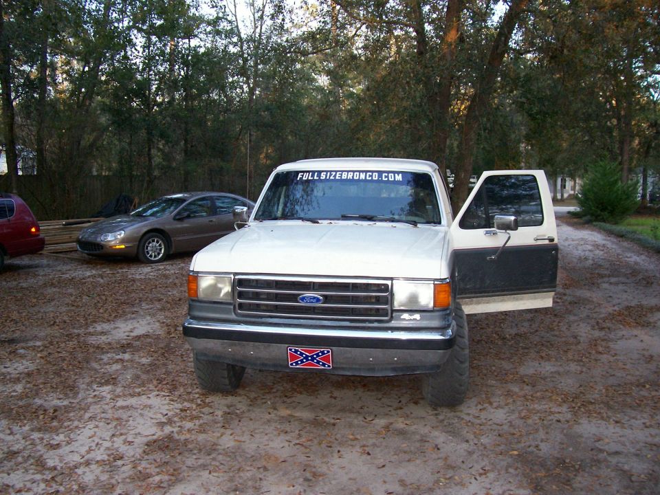 White Ford Bronco front view with open driver door, FULLSIZEBRONCO.COM windshield banner, parked on dirt driveway
