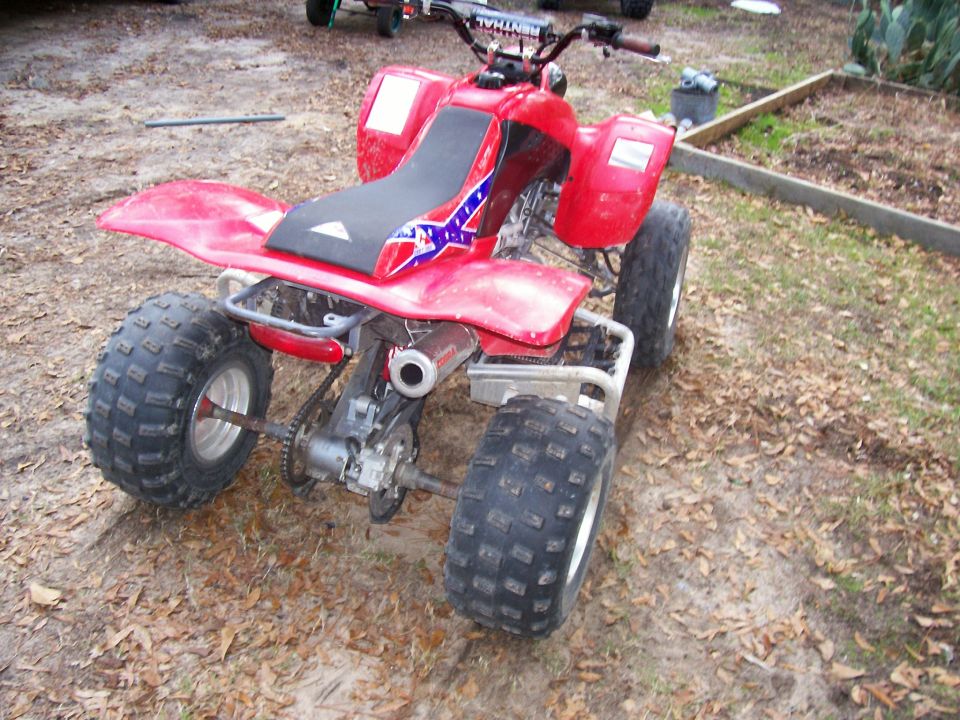 Red sport ATV quad parked on dirt, rear 3/4 view with black knobby tires.