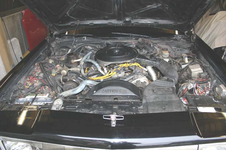 Engine bay of a black sedan with round air cleaner, yellow plug wires, and CAUTION FAN shroud