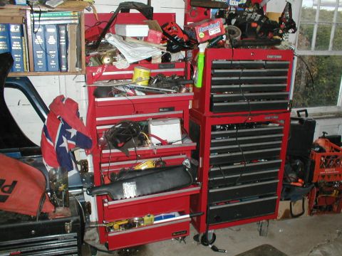 1979 Ford Bronco - A cluttered garage scene with multiple red tool chests and various car parts, including a car door with a flag.