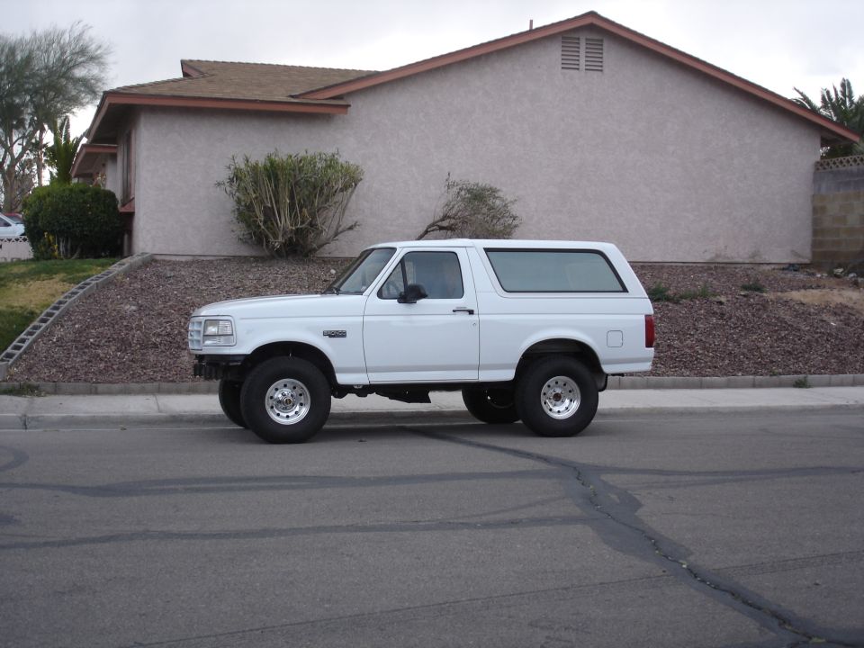 White Ford Bronco SUV, side view, lifted with chrome steel wheels, parked on street by tan house
