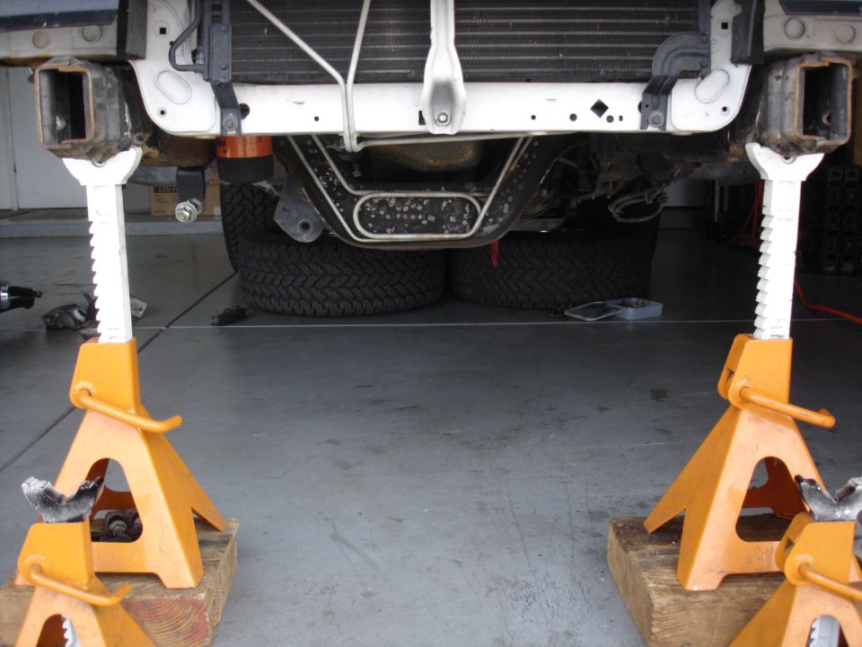 Truck front raised on two yellow jack stands in garage, dual rear tires and radiator visible underneath