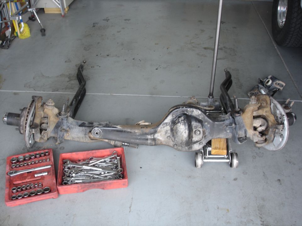 Removed front axle assembly with brakes on a floor jack in a garage, with socket set and wrenches in front