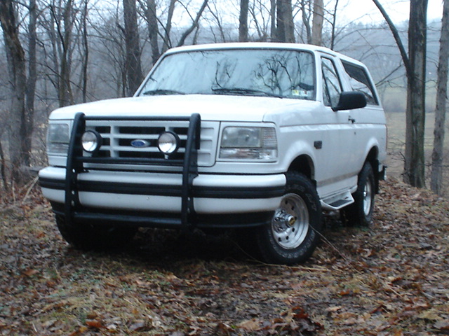 White 1995 Ford Bronco with bull bar parked on a dirt road in woods