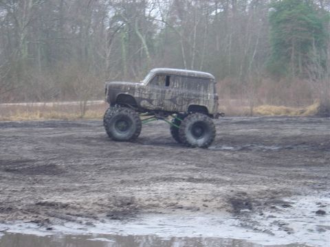 1987 Suzuki Samurai - Custom monster truck with large tires navigating a muddy terrain with trees in the background.