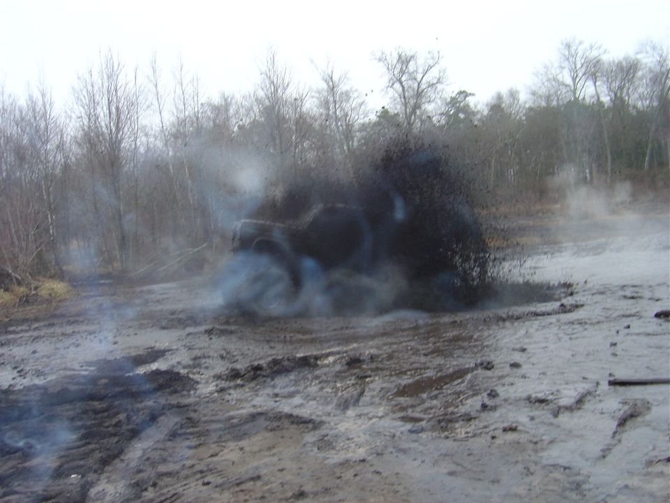Dark off-road vehicle splashing mud and emitting smoke while driving through a muddy terrain with trees in the background
