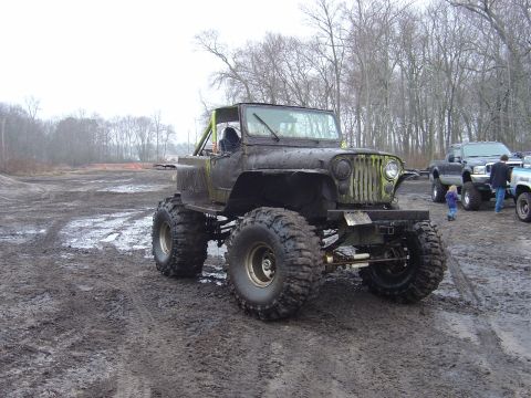1987 Suzuki Samurai - Muddy off-road Jeep CJ-5 with oversized tires and custom roll cage, parked in a muddy field.