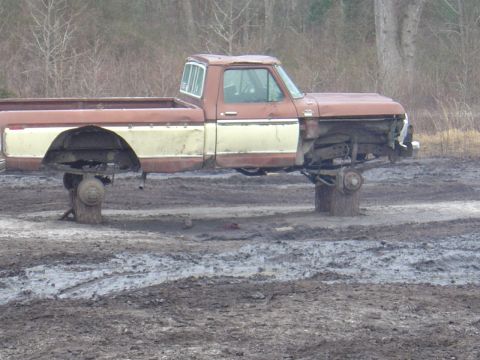 1987 Suzuki Samurai - A weathered pickup truck sits elevated on blocks in a muddy field, lacking its wheels and front bumper.