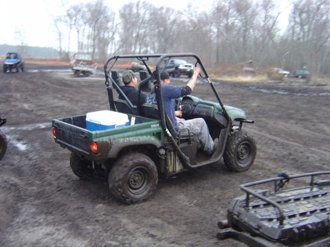 1987 Suzuki Samurai - Two people ride in a green Yamaha utility vehicle through a muddy off-road area. Other vehicles are present.