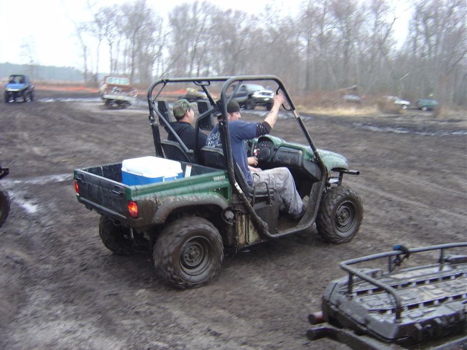 Green Yamaha utility vehicle with two occupants driving on a muddy track. Another vehicle is visible in the background