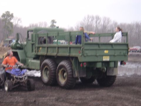 1987 Suzuki Samurai - A large green military truck is seen from the rear with passengers in the cargo bed and an ATV nearby.