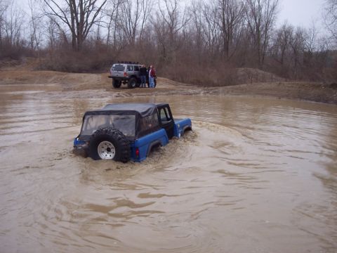 1974 Ford Bronco - Two off-road vehicles navigate a muddy terrain, with one vehicle partially submerged in water.