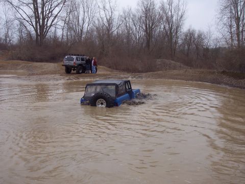1974 Ford Bronco - A blue Jeep Wrangler is stuck in a muddy puddle while another SUV waits on the bank.