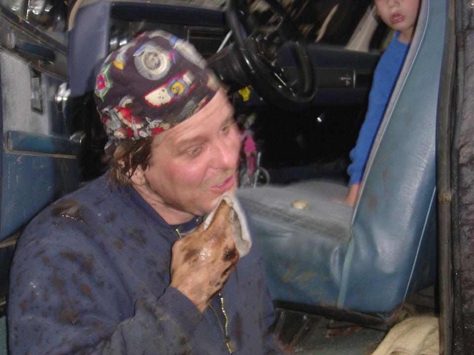 Person with dirty hands wiping face with a cloth inside a vehicle, with a child in the background