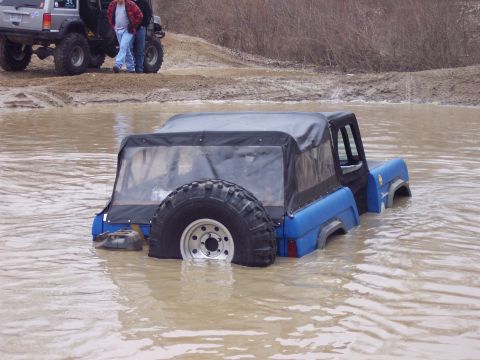 1974 Ford Bronco - This blue off-road vehicle is partially submerged in murky water, with other vehicles visible on the muddy bank.