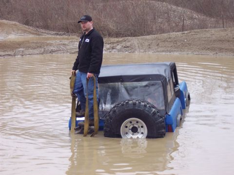 1974 Ford Bronco - A man stands on the side of a blue off-road vehicle stuck deep in muddy water, holding tow straps.