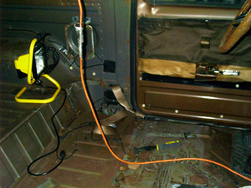 Interior of a vehicle with stripped floor, door panel, and steering wheel visible. A work light and orange extension cord are present