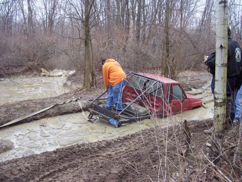 1974 Ford Bronco - A red SUV with a roll cage is stuck deep in the mud while people attempt to free it.