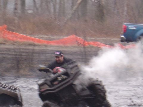 1987 Suzuki Samurai - A man navigates an ATV through a muddy track, with a blue pickup truck visible in the background.