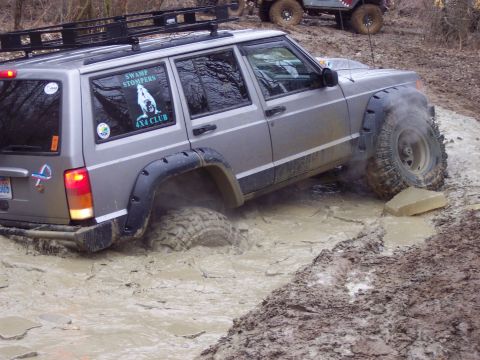 1974 Ford Bronco - A silver Jeep Cherokee XJ navigates a muddy obstacle course, with another off-road vehicle visible in the background.