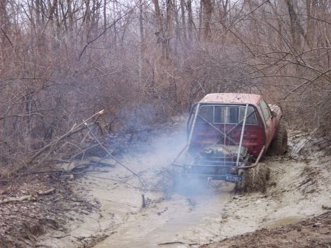 1974 Ford Bronco - A modified pickup truck navigates a challenging muddy path surrounded by trees.