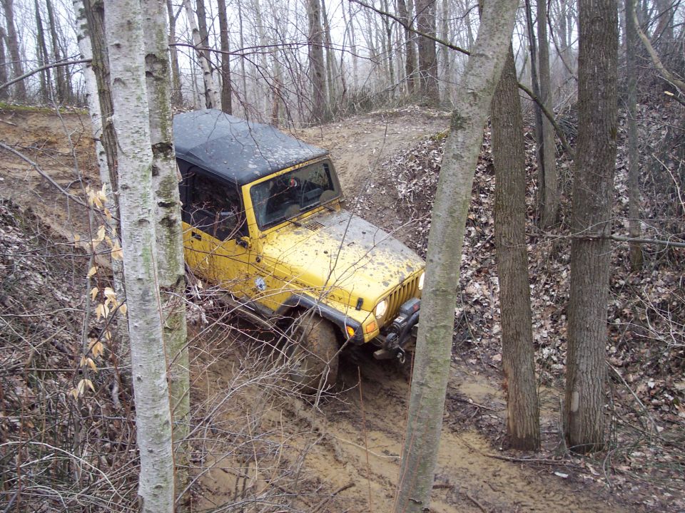 Yellow Jeep Wrangler on a muddy trail, front-right tire stuck in mud, trees and branches surrounding