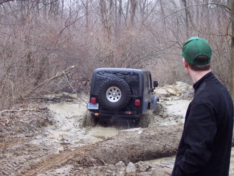 1974 Ford Bronco - A Jeep Wrangler navigates a challenging muddy path surrounded by trees.