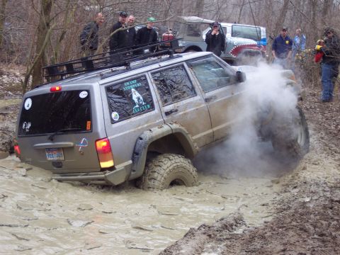 1974 Ford Bronco - A silver Jeep Cherokee XJ navigates a muddy obstacle, kicking up smoke, while other off-road vehicles and spectators watch.