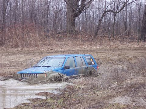 1974 Ford Bronco - A blue Jeep Grand Cherokee is stuck in a muddy puddle in a wooded area.