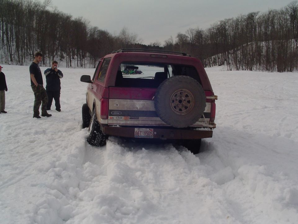 Red Ford Bronco stuck in snow, spare tire on back. People standing around