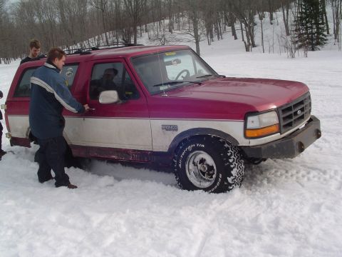 1994 Ford Bronco - A red and white Ford Bronco is stuck in deep snow with two people attempting to get it out.
