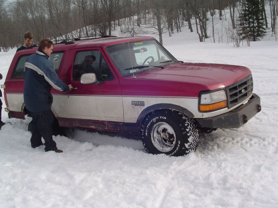 Red and white Ford Bronco stuck in snow, driver's door open, two people nearby
