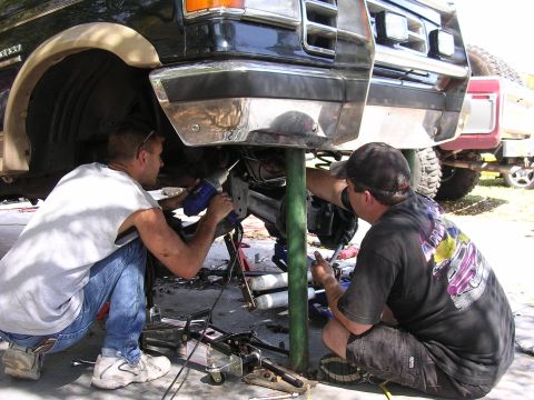 1988 Ford Bronco - Two men perform suspension work on a truck, with a second vehicle visible behind it.
