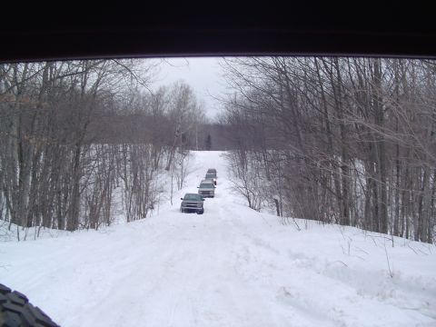 1994 Ford Bronco - Several SUVs travel in a convoy up a snowy, tree-lined road under an overcast sky.