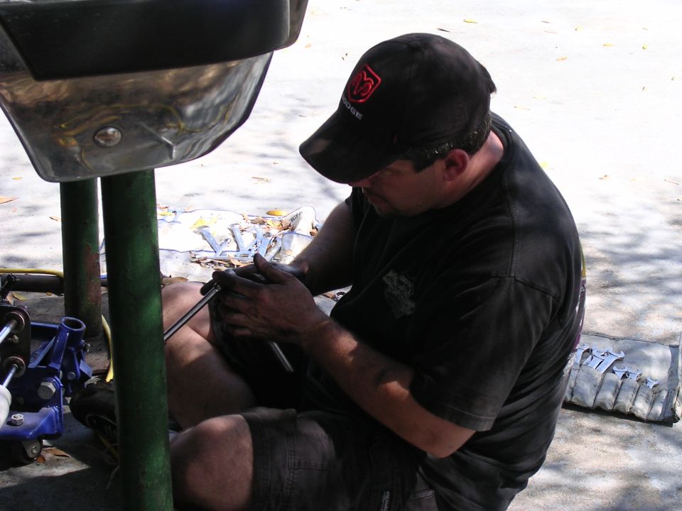 Man in black t-shirt and cap works on a metal vehicle part with a socket wrench