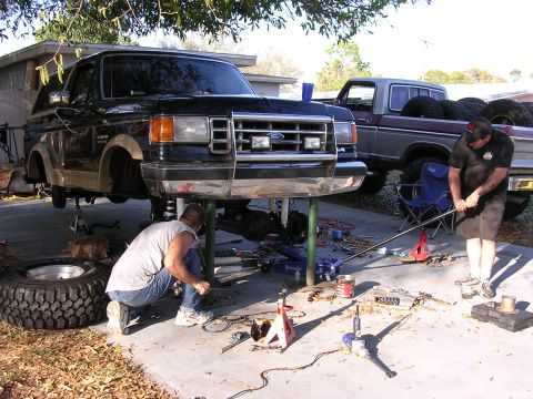 1988 Ford Bronco - Two men work on a lifted black Ford Bronco and a pickup truck in a driveway.