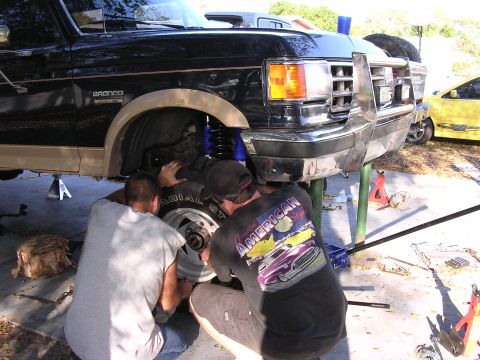 1988 Ford Bronco - Two men work on the front suspension of a black Ford Bronco, which is raised on jack stands.