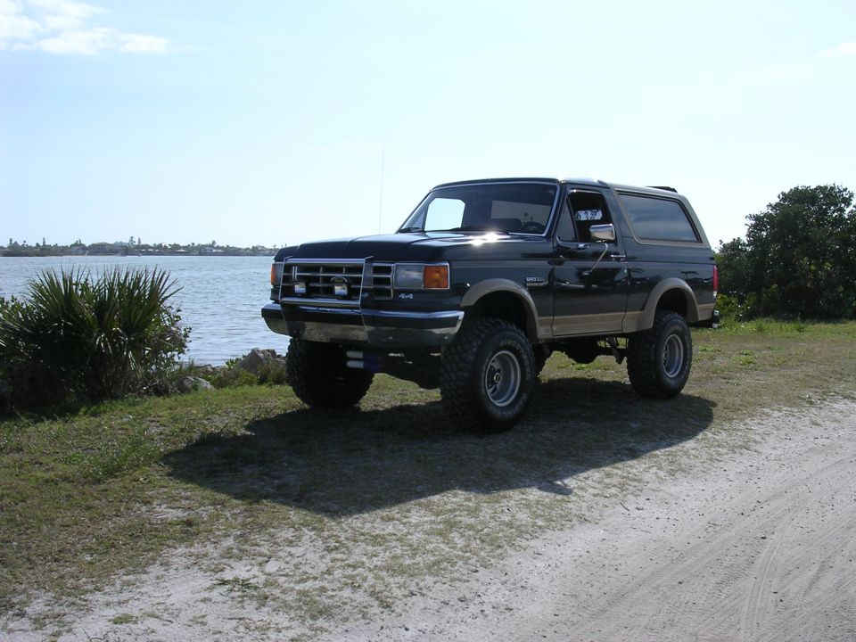 Black lifted Ford Bronco parked on a dirt road near water, with tan side trim and chrome wheels