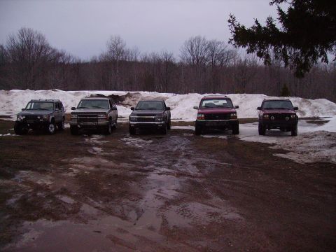 2000 Chevrolet Tahoe - A lineup of five SUVs are parked in a muddy lot with snow-covered ground and trees in the background.